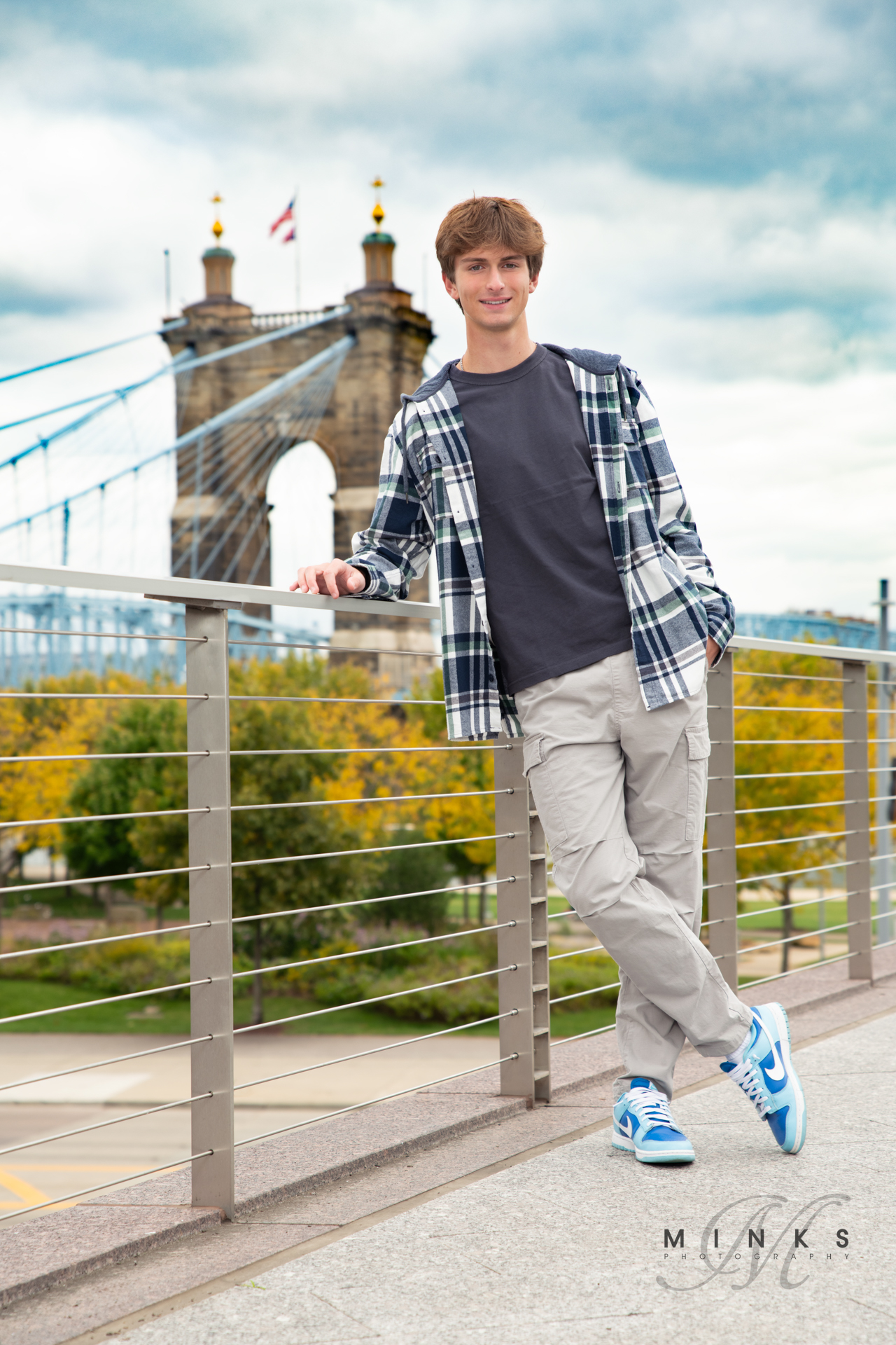 high school senior guy leaning on a rail with the Roebling bridge behind him